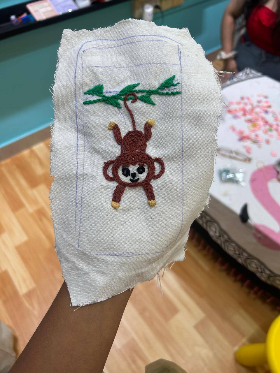 Hand holding embroidery fabric with a stitched hanging monkey design on a branch.