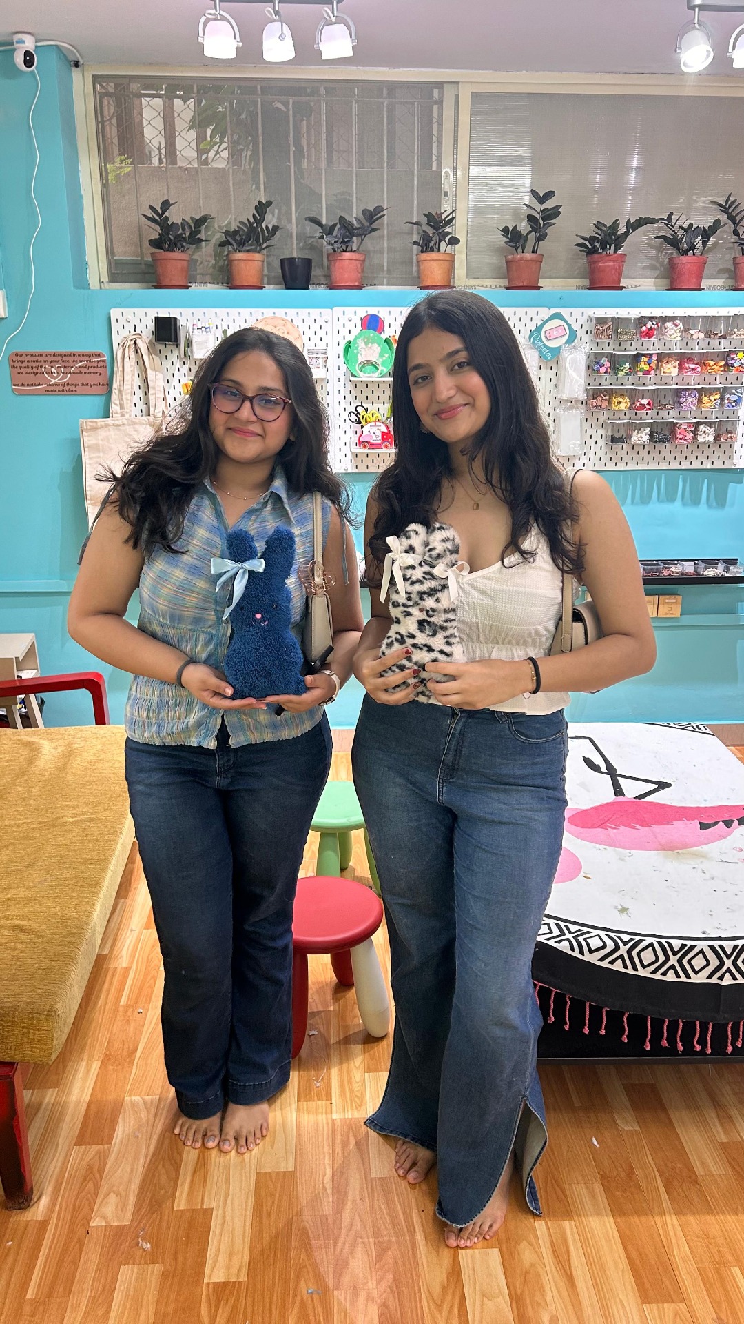 Two women standing barefoot holding finished plush bunny toys in a creative workspace.