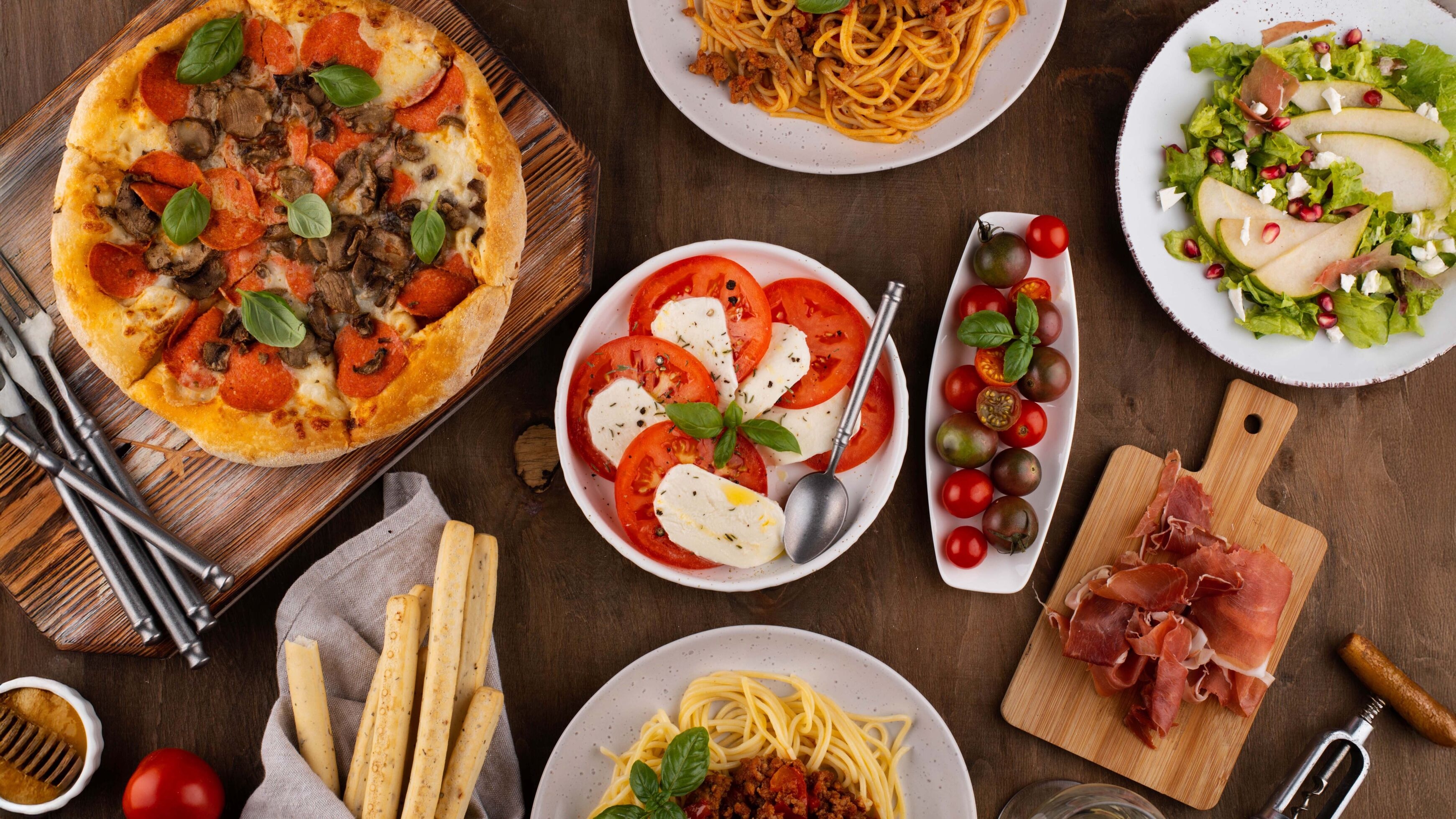 Authentic Italian spread with pizza, spaghetti, caprese salad, mixed greens, cured ham, and breadsticks on a wooden table.