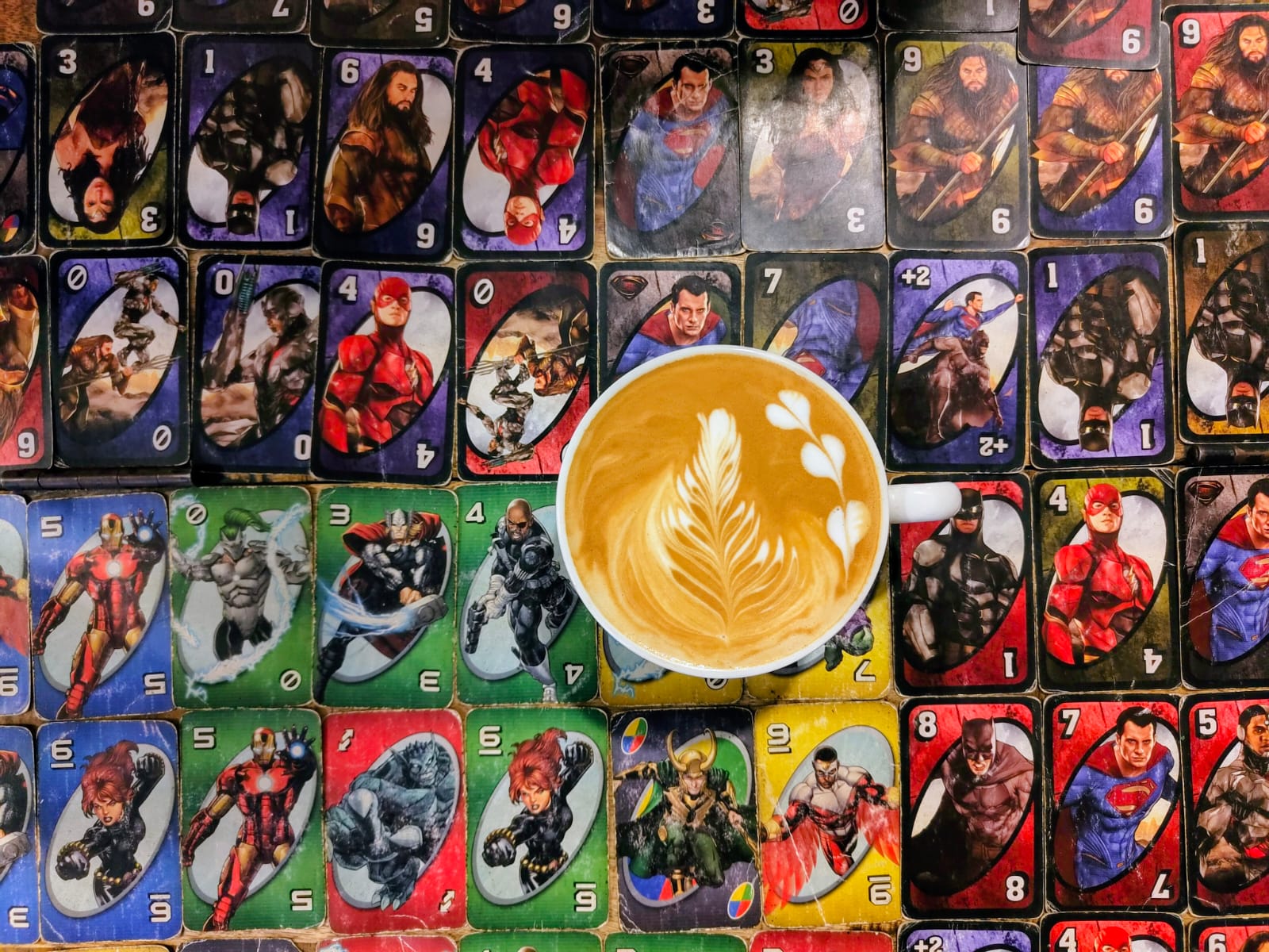 Cup of latte with leaf and heart latte art placed on a table covered with superhero themed playing cards.