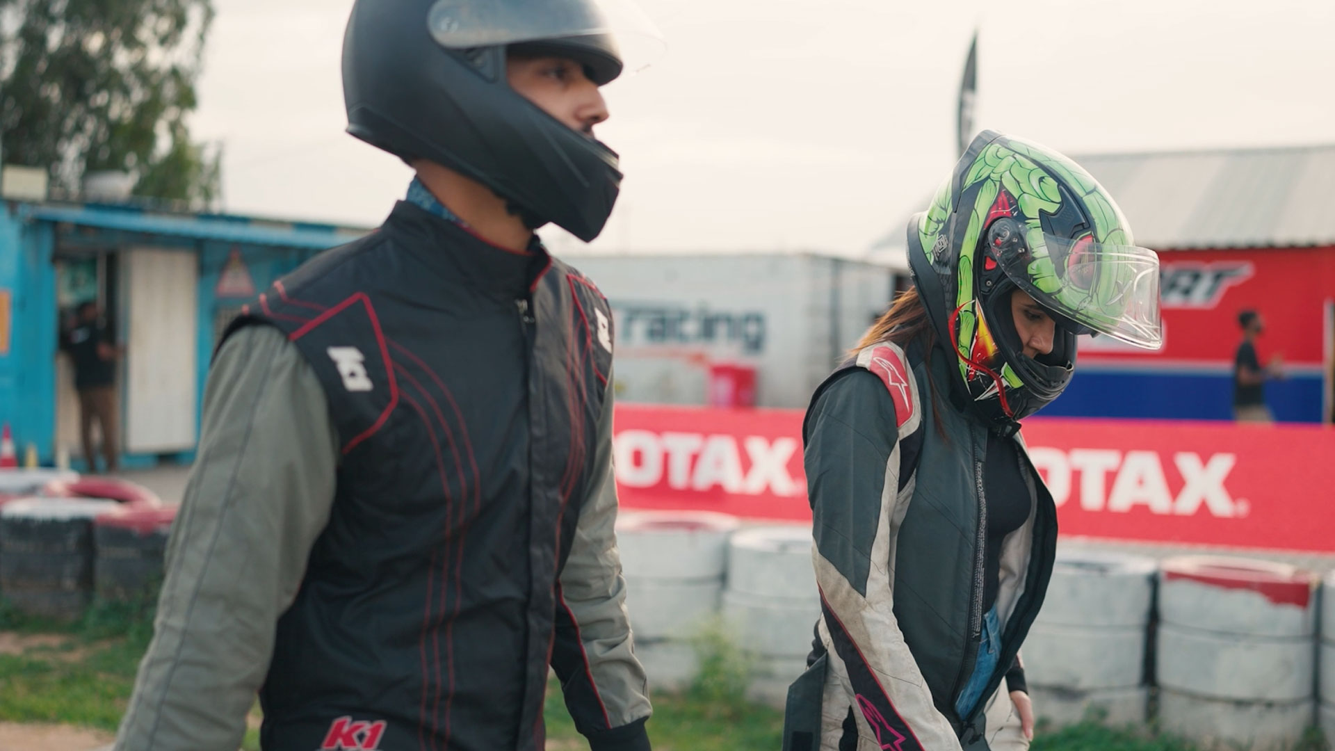Two individuals in racing gear and helmets walking towards the track.