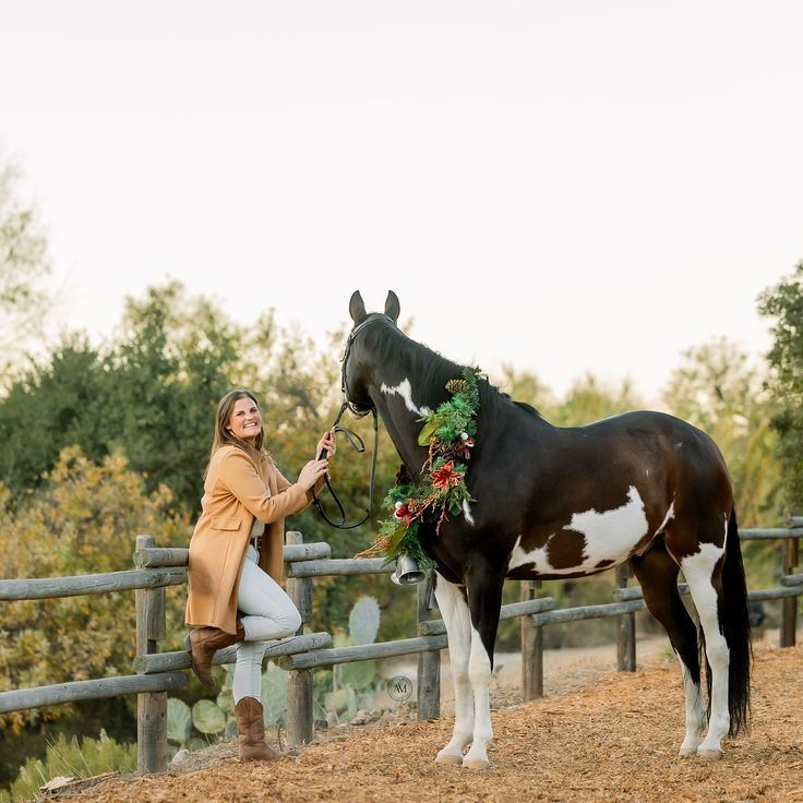 Smiling woman holding reins of a black and white horse decorated with a Christmas wreath outdoors.