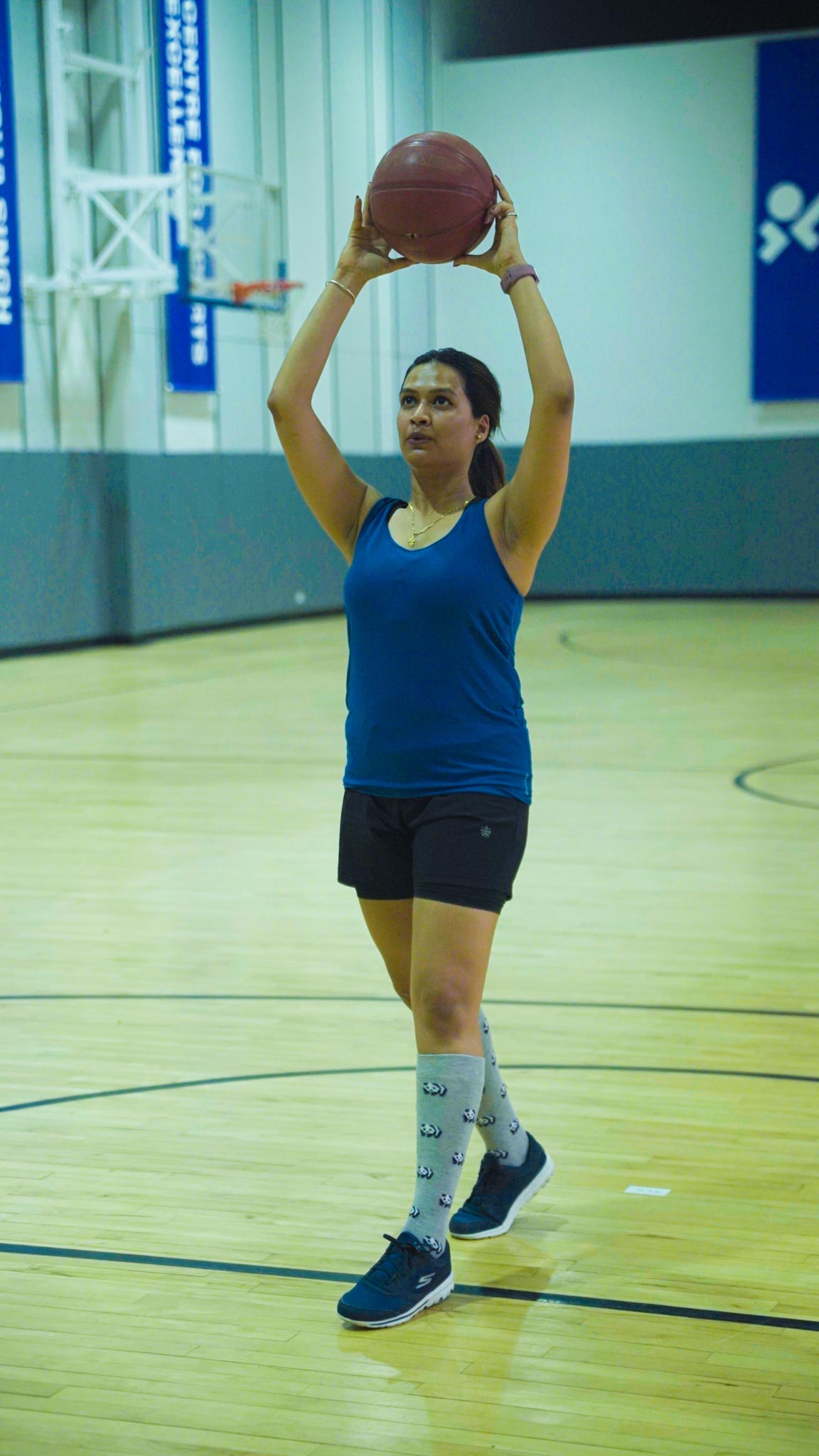 A woman in blue top lifting a basketball overhead for a shot in an indoor Bengaluru basketball court.