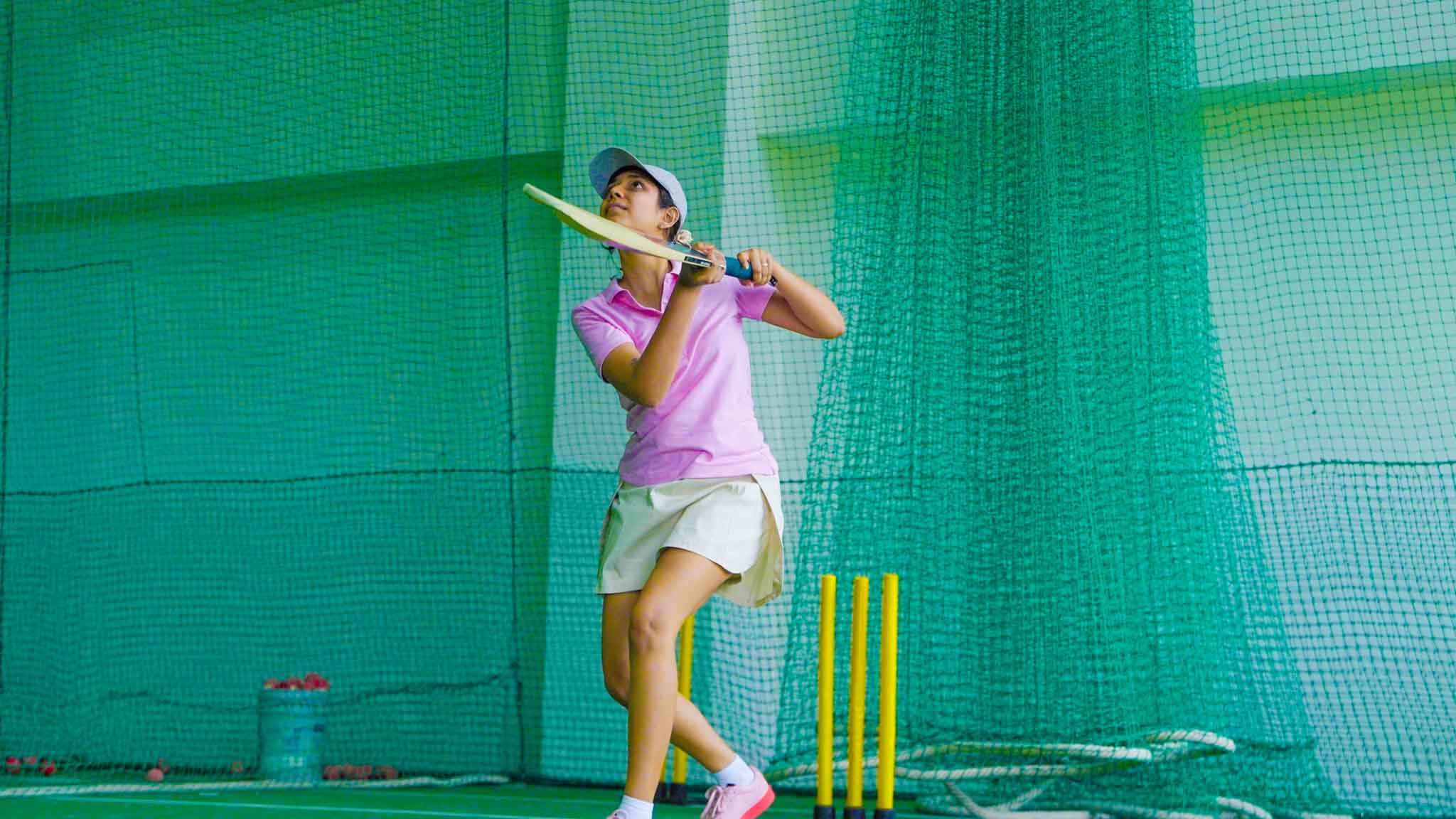 Woman batting in cricket net practice wearing cap and sportswear.