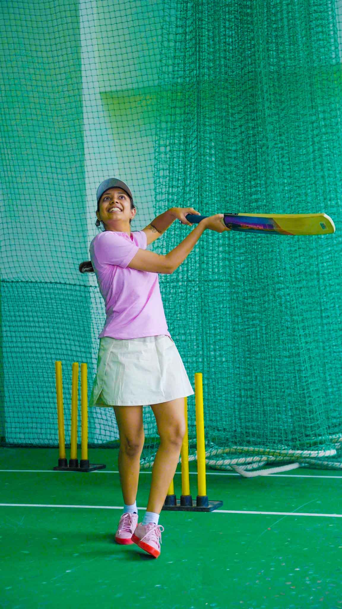 Woman in pink shirt and cap batting in an indoor cricket net in Bengaluru.