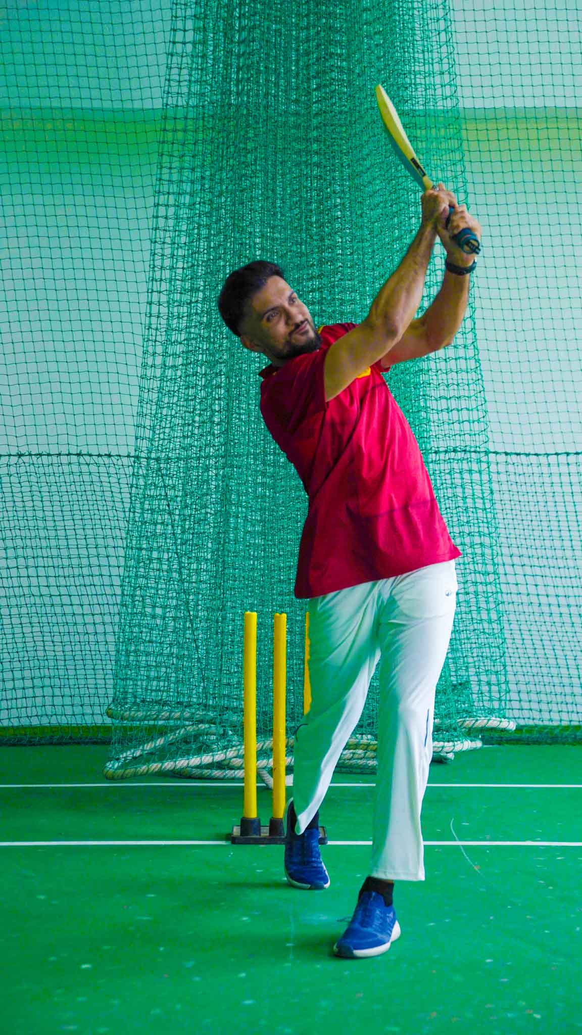 Man following through on a cricket shot with bat raised indoor cricket arena.