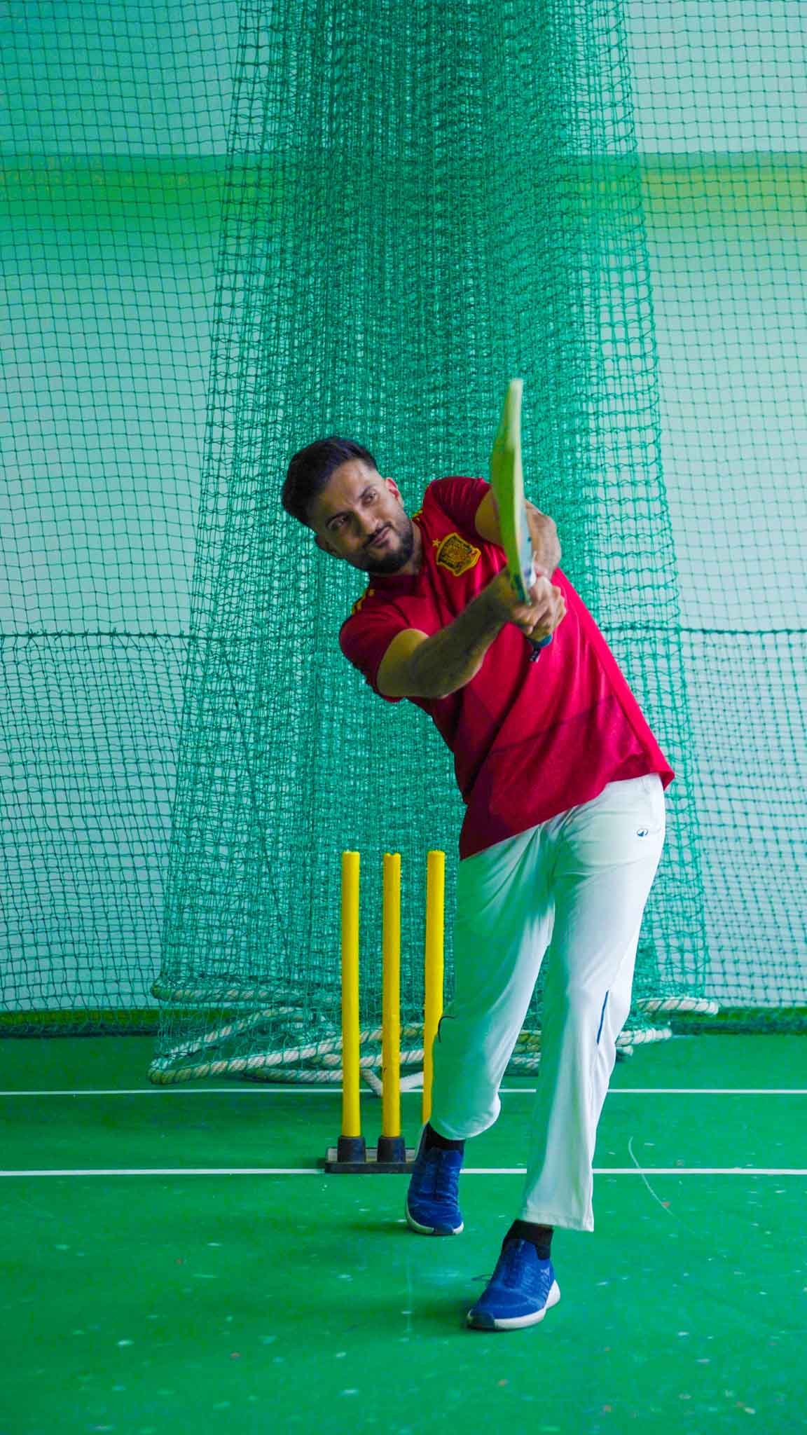 Man in red shirt playing a cricket shot inside an indoor net practice facility in Bengaluru.