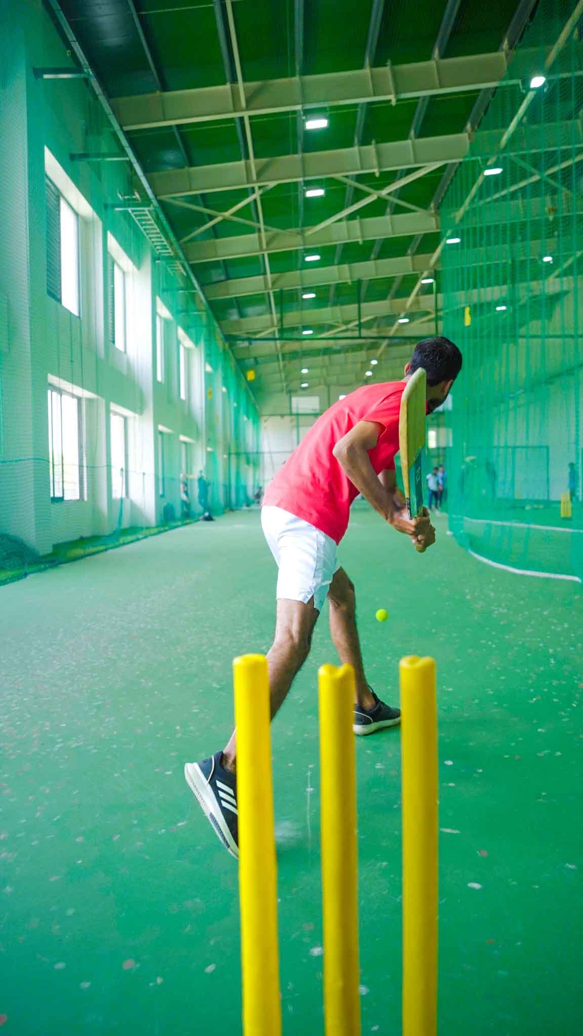Man in red shirt pitching cricket ball in an indoor cricket coaching session.