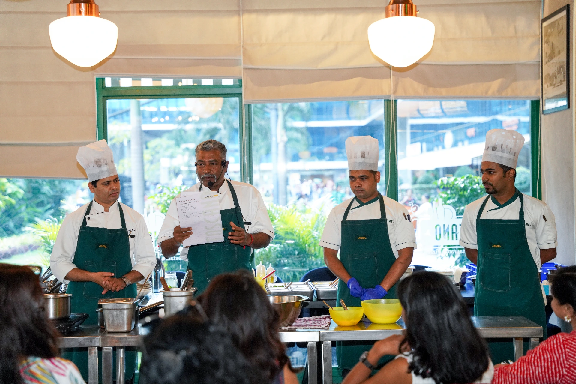 Chefs in white uniforms and green aprons conducting a gnocchi cooking demo in front of an audience.