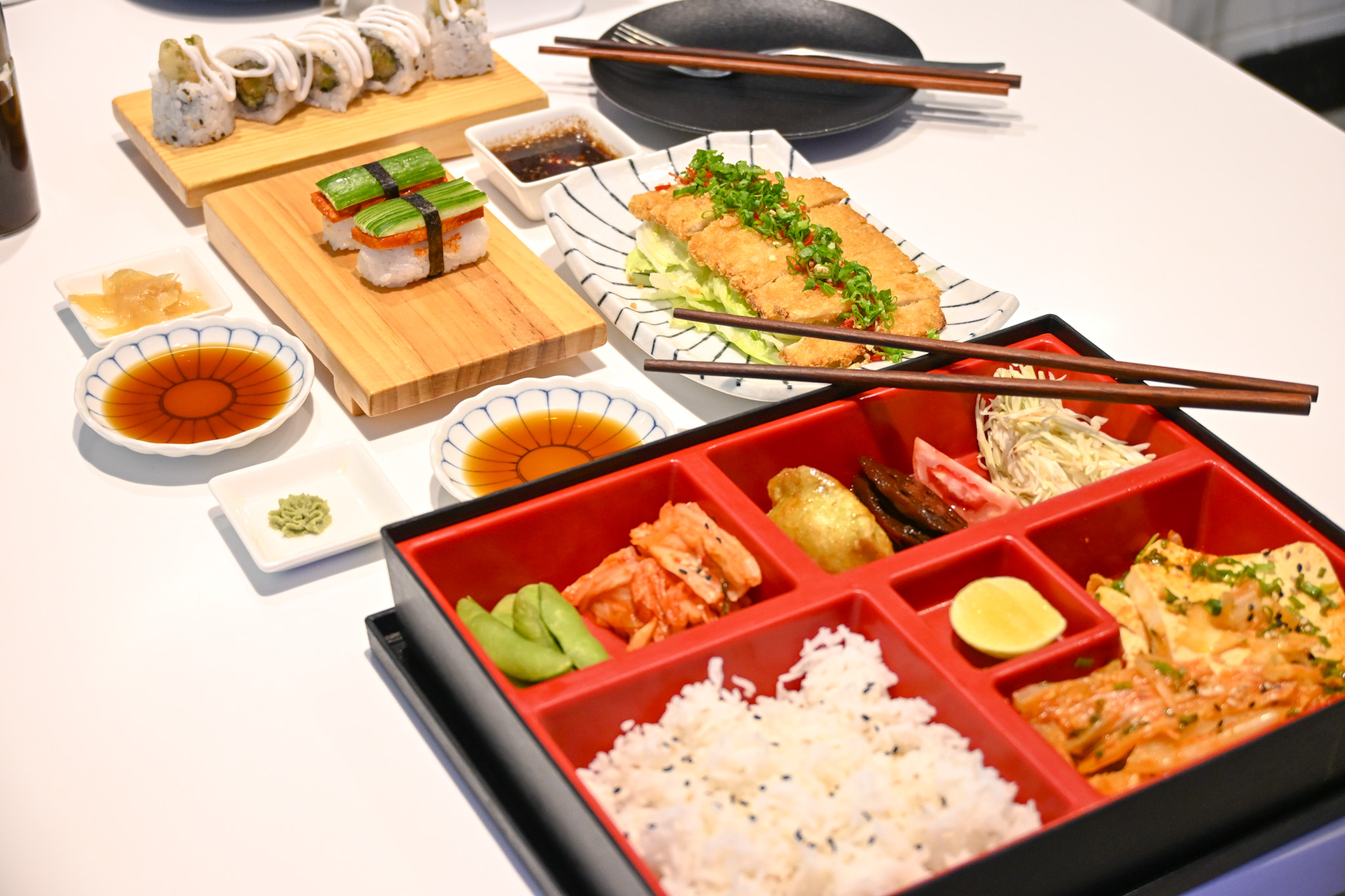 Japanese meal spread on white table with sushi, bento box, dipping sauce and chopsticks.