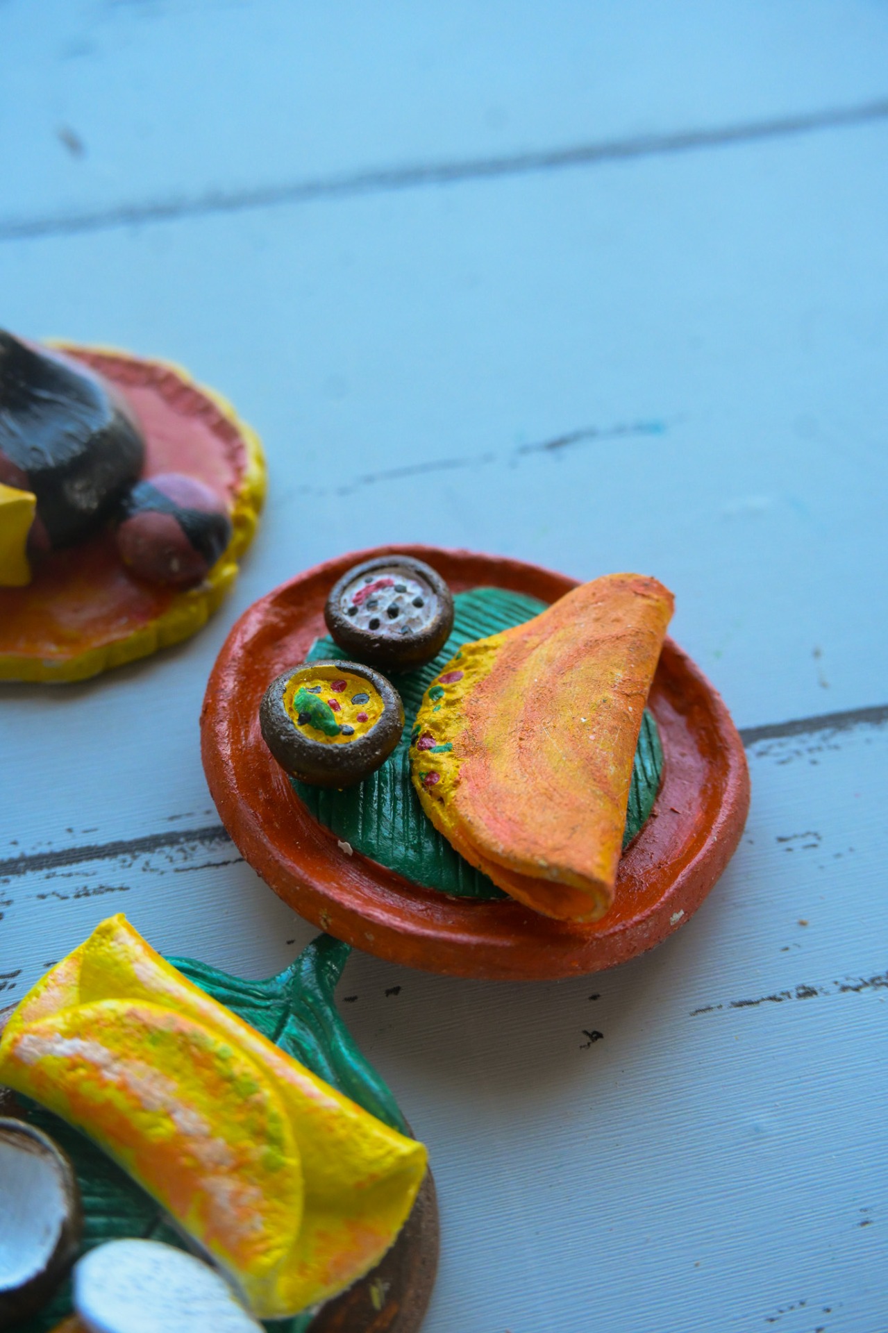 Miniature magnet art showing a traditional Indian snack plate with folded dosa and two small bowls on a leaf on a clay plate.