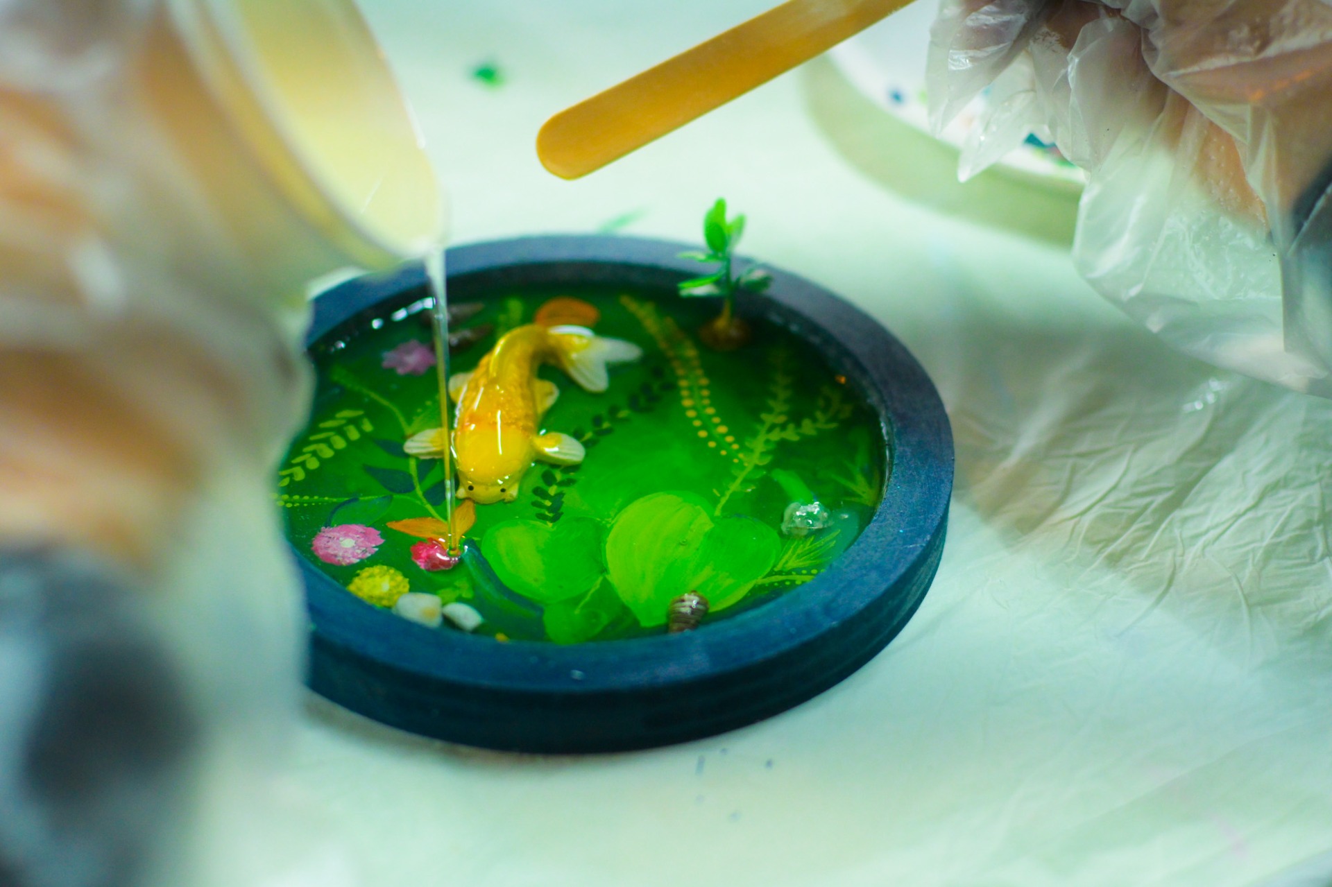 Pouring clear resin over a decorated resin tray with painted fishes and plants.