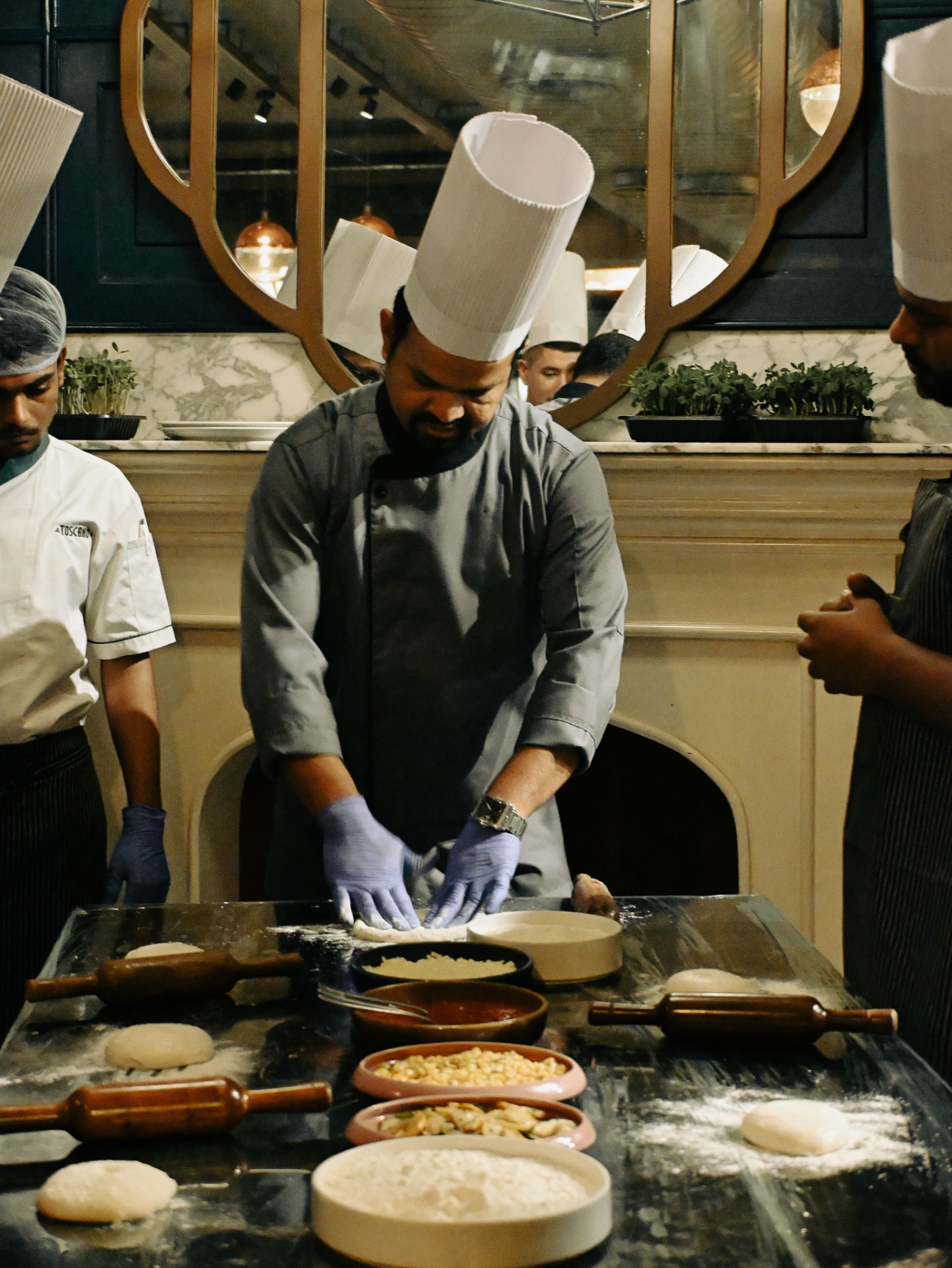 Chef in uniform demonstrating pizza dough preparation at a cooking class table with ingredients arranged.