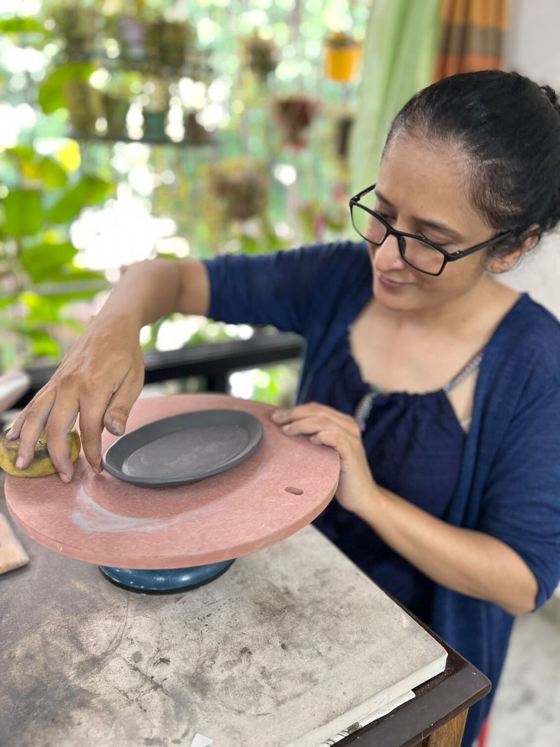 Woman with glasses smoothing a clay plate on a pottery wheel in a bright room with plants.