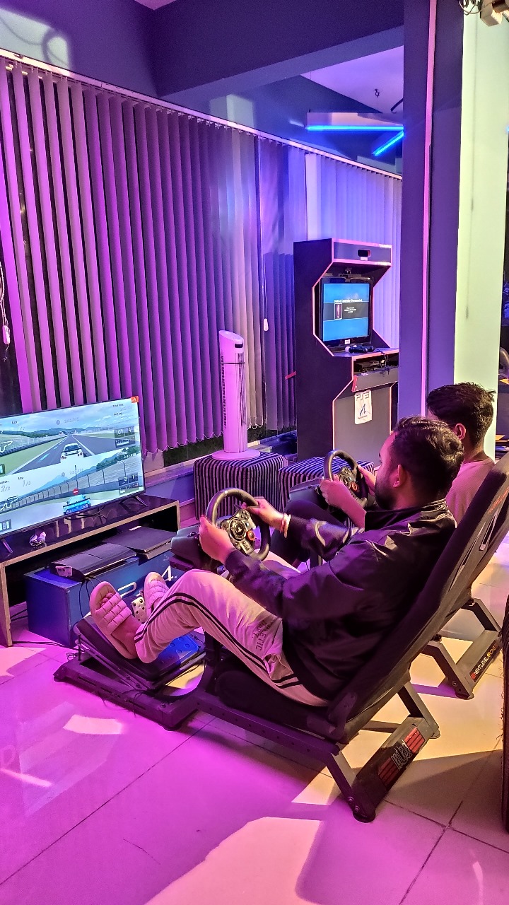 Two players racing on car racing simulators using steering wheels and pedals with a large TV displaying the racing game.