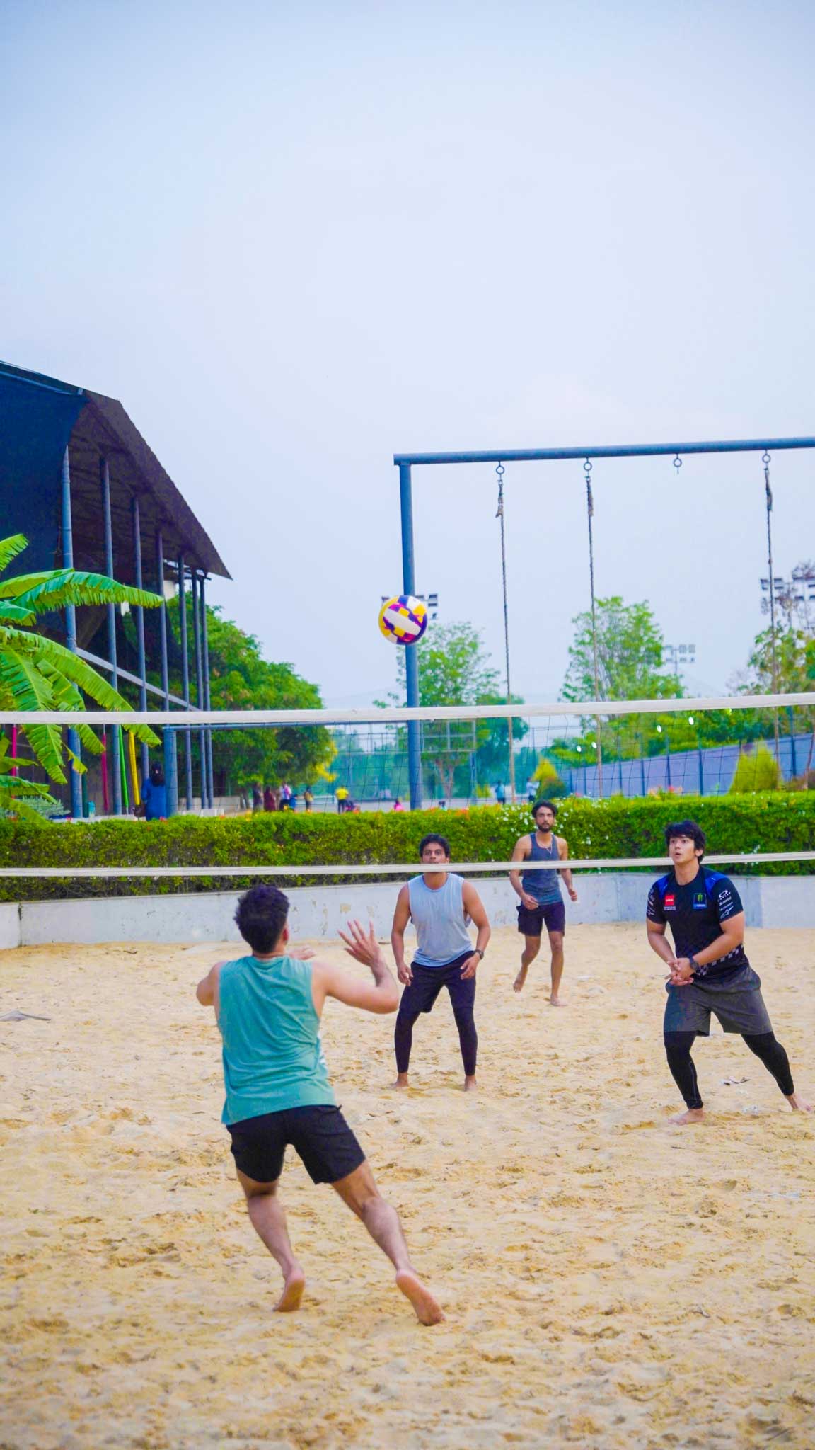 Group playing beach volleyball on a sand court with net and trees in background.