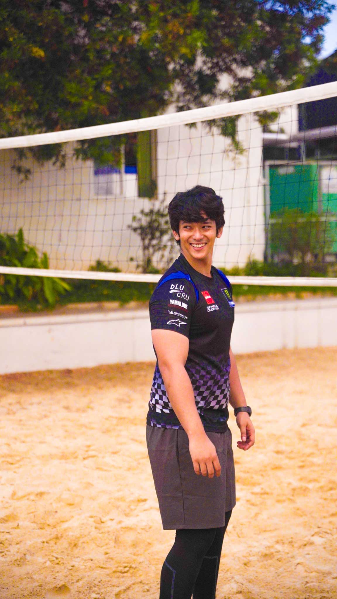 Man smiling and standing on the sand volleyball court near the net with trees around.