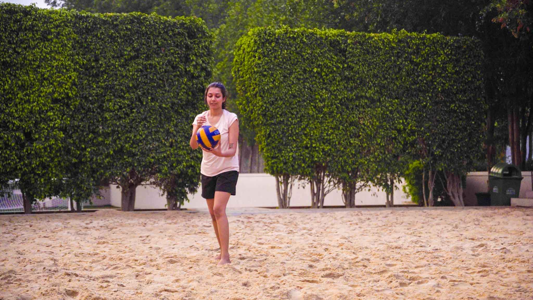 Woman in casual sportswear holding a volleyball on the sandy court surrounded by greenery.
