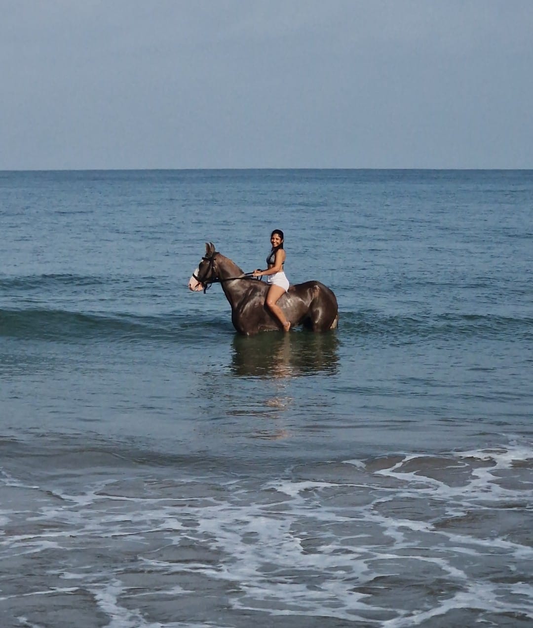 Woman riding horse waist-deep in ocean water during beach surf with horses experience.