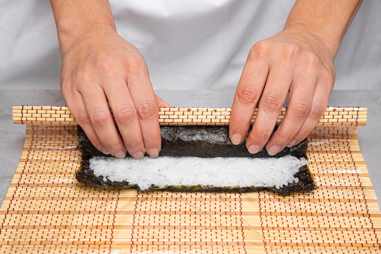 Close-up of hands rolling sushi on a bamboo sushi mat, showing the rice and nori sheet.