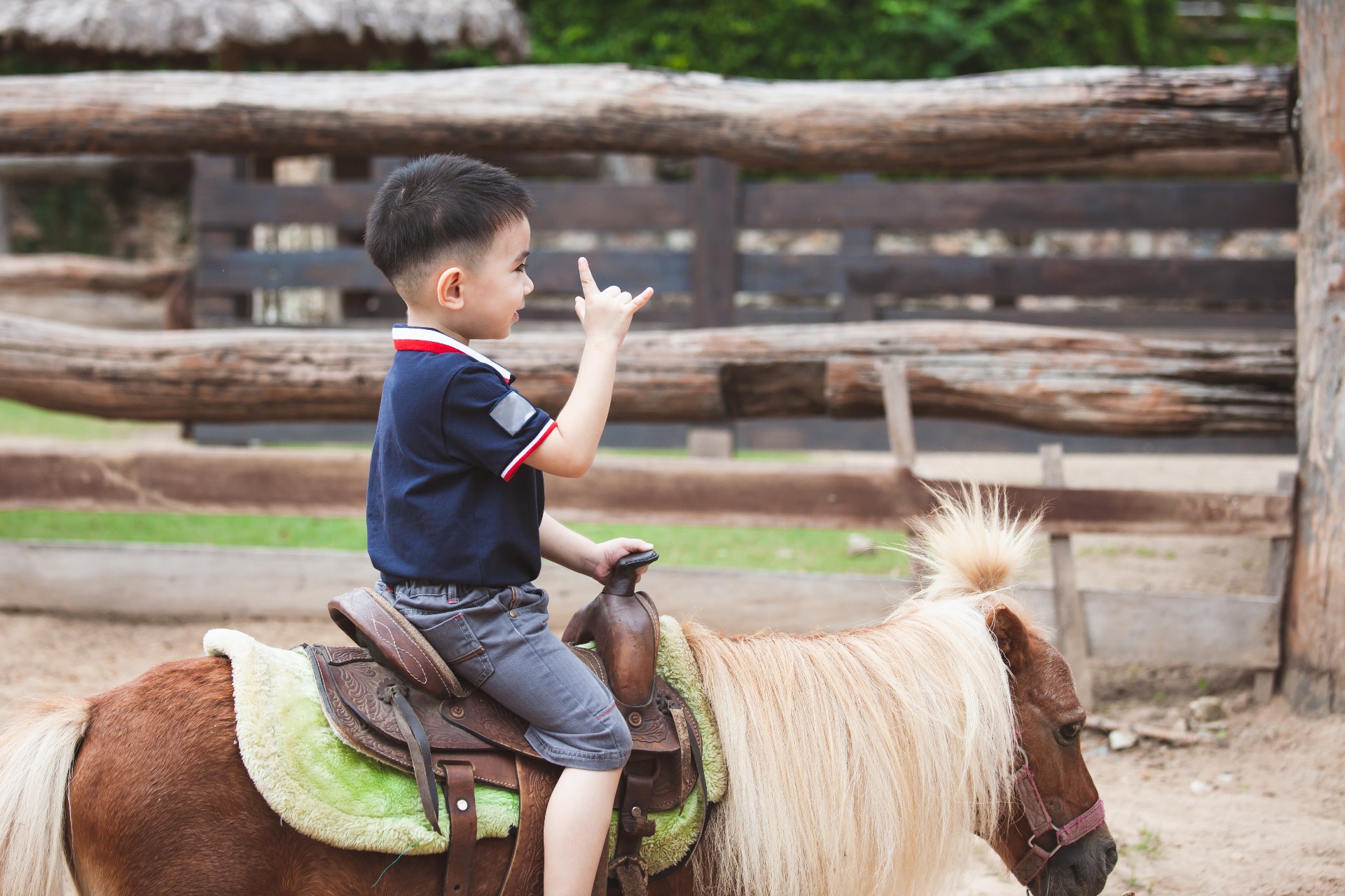 Young boy making a playful hand gesture while riding a tan pony at a farm.