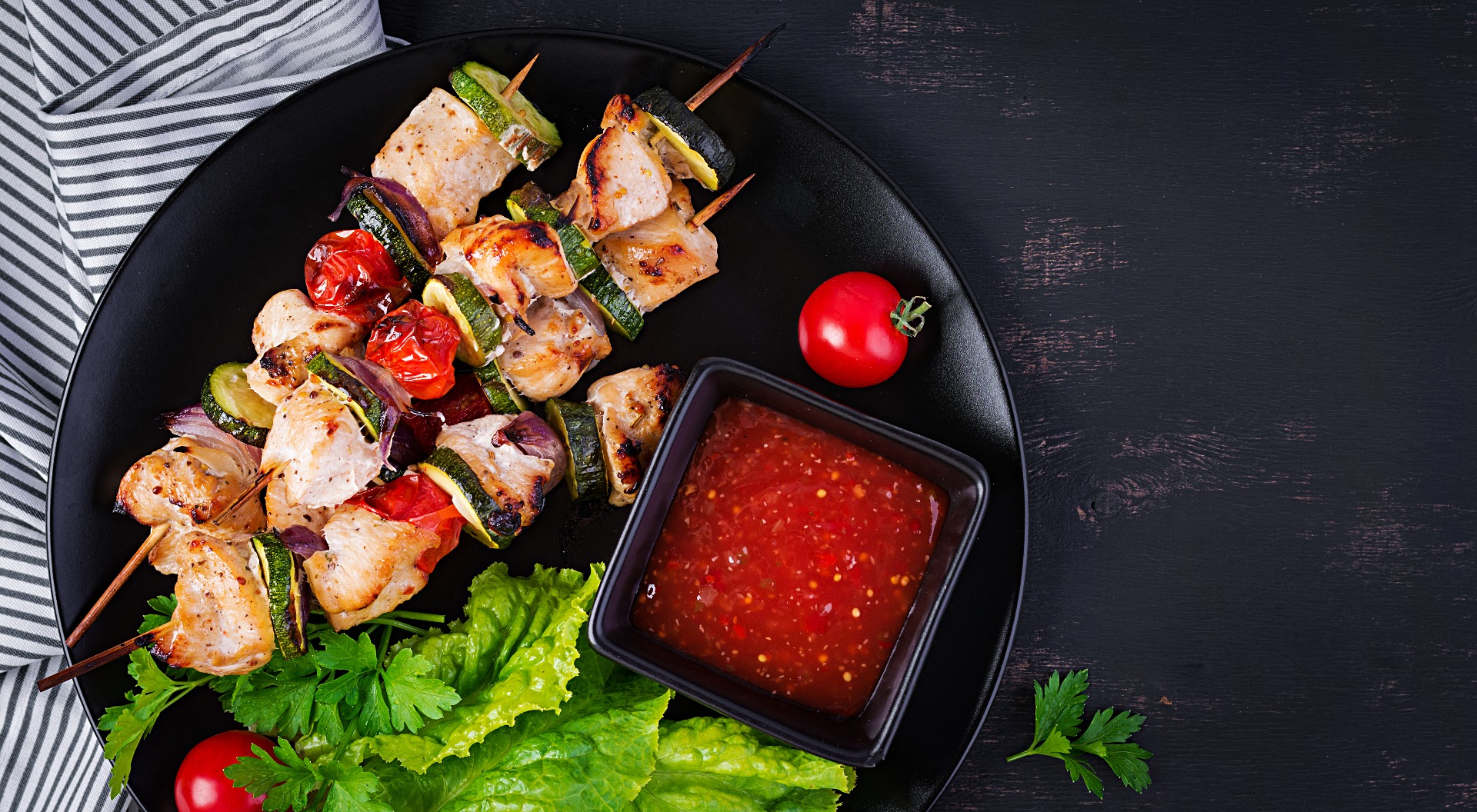 Chicken shish kebabs with zucchini, tomatoes and onions served on a plate with dipping sauce.
