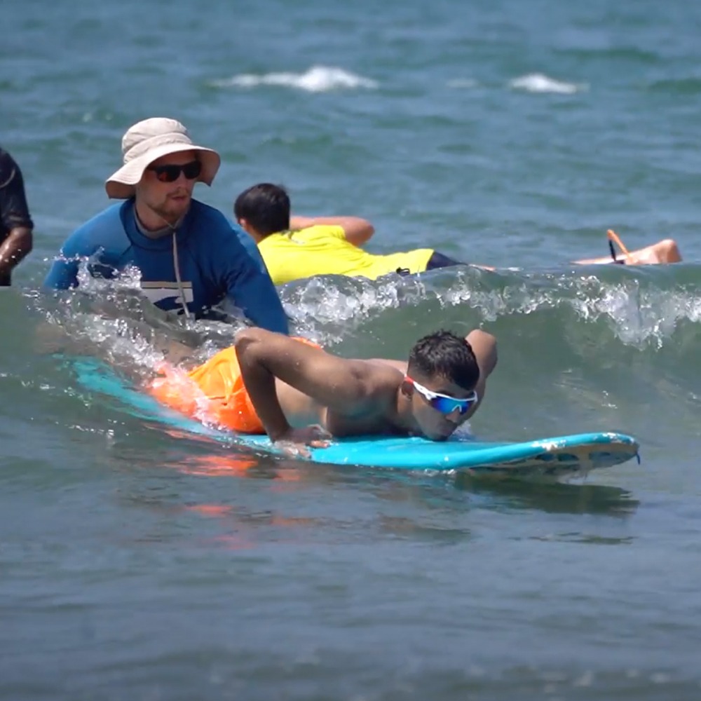 Instructor assisting a beginner to paddle on a surfboard in the water during surf lesson.