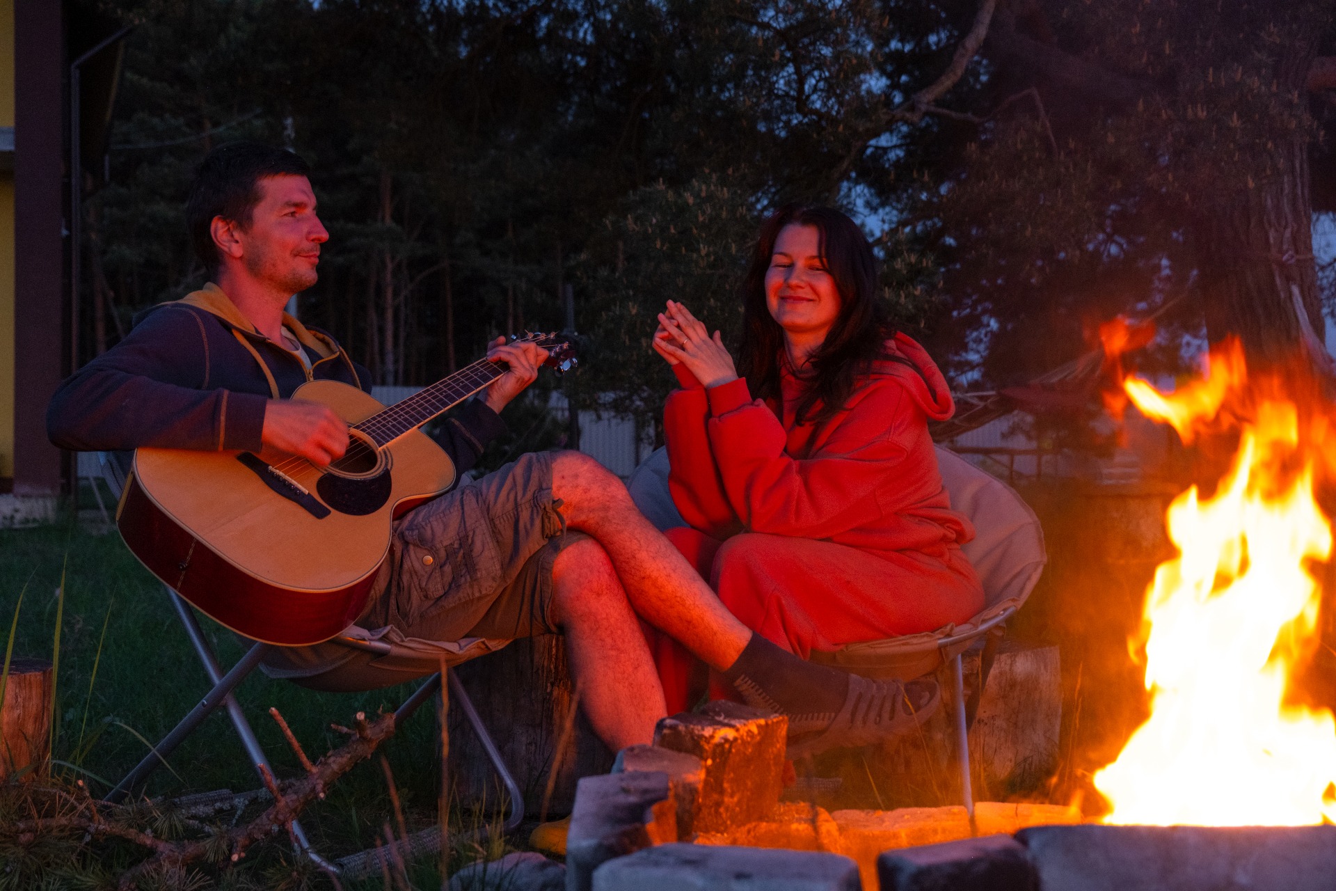 Man playing guitar while woman sings and claps near a bonfire at night.