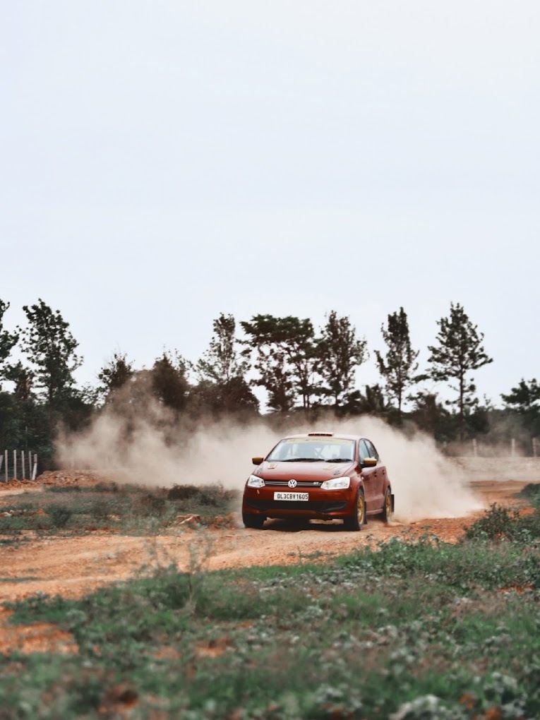 Red rally car kicking dust behind as it races on a countryside dirt rally track.